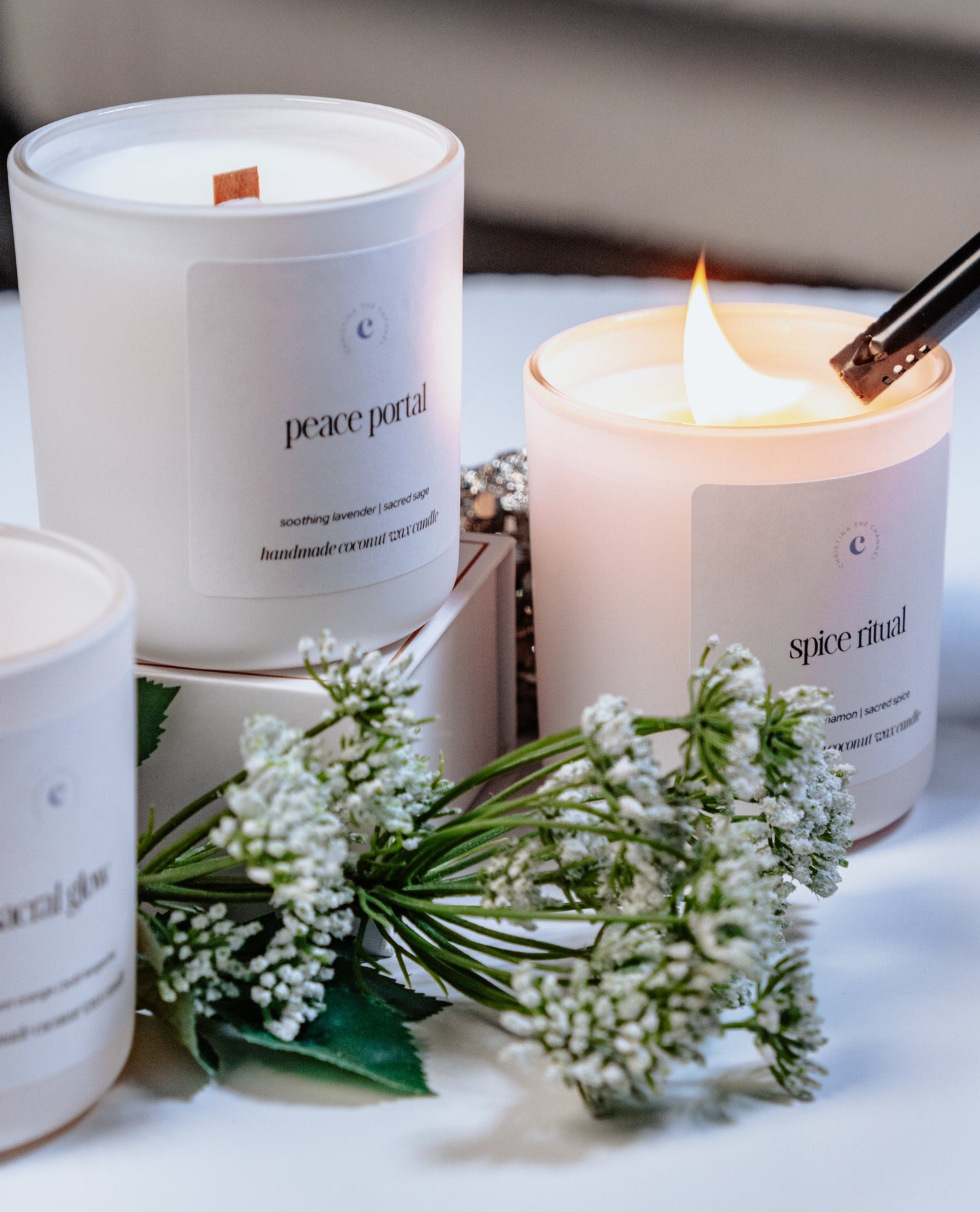 Three candles with labels on a white surface, surrounded by small white flowers.