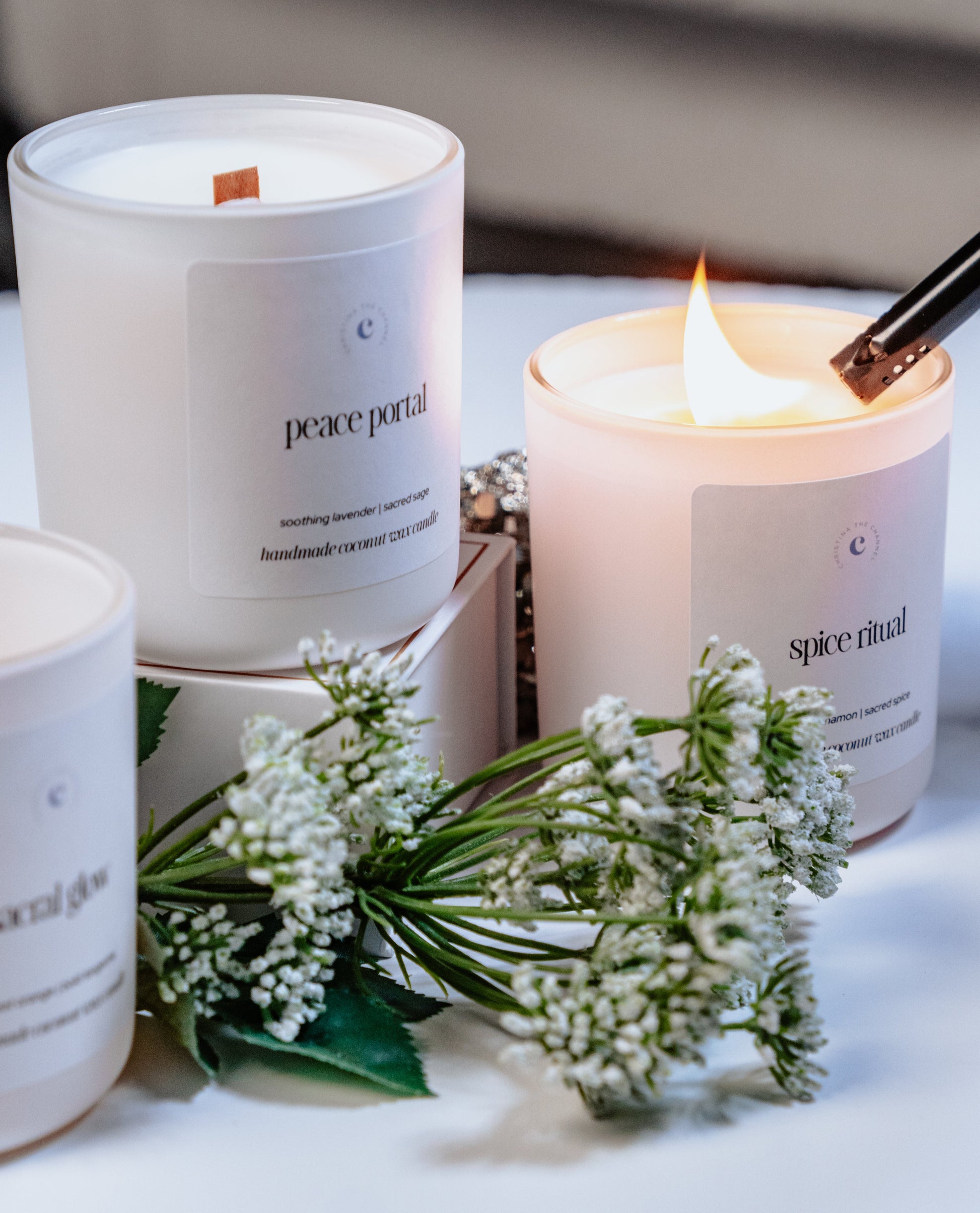 Three candles with labels on a white surface, surrounded by small white flowers.